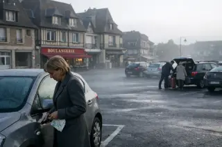 Morning mist over a quiet Deauville parking lot, woman with city map locking her car, townhouses and a boulangerie visible, subdued natural light.
